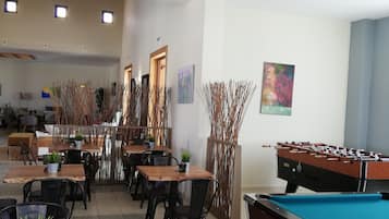 Una stanza con tavolo da biliardo, tavoli e sedie di legno e piante in vaso.