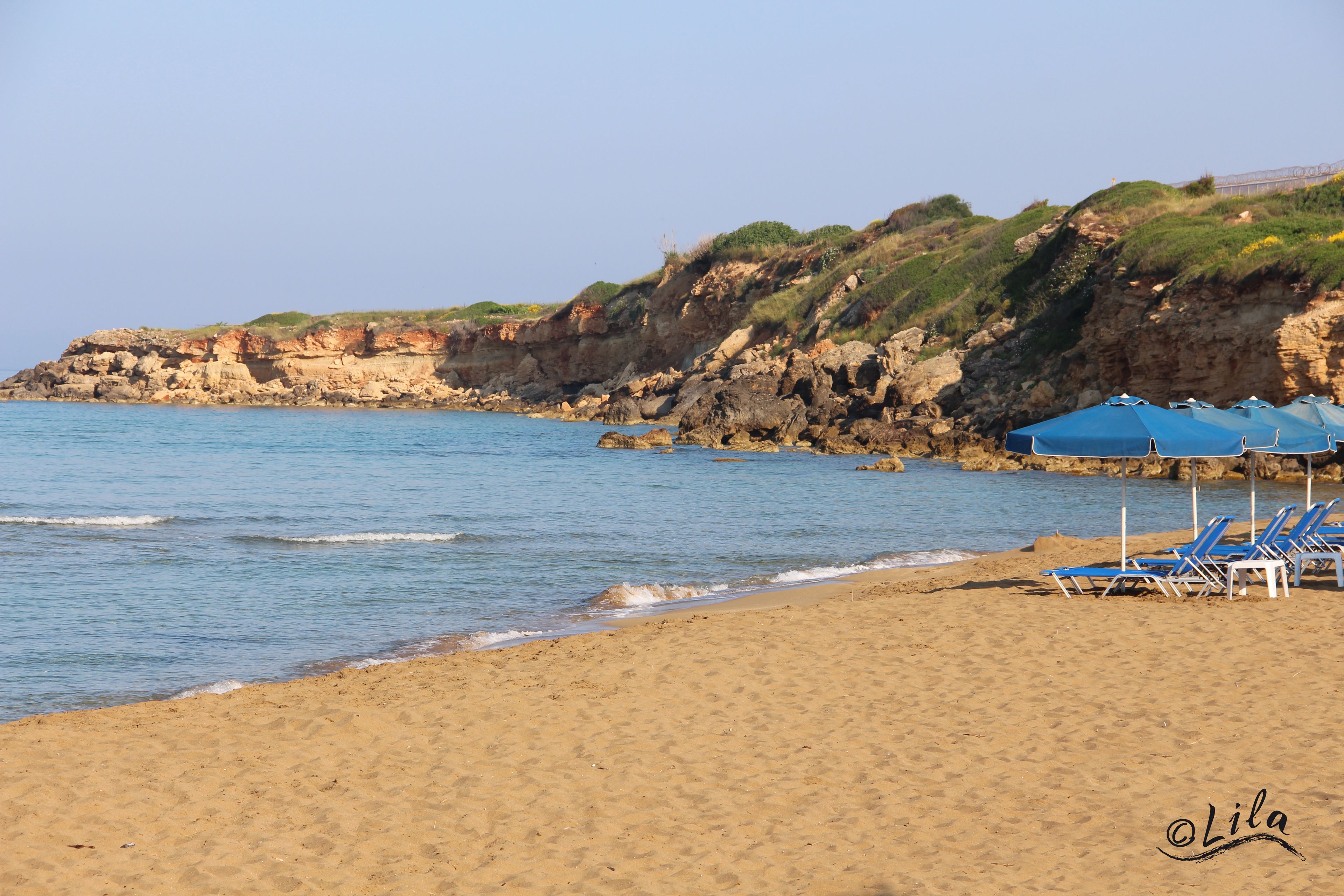 Una spiaggia con acque cristalline, una riva sabbiosa e scogliere rocciose. Si vedono ombrelloni e lettini blu.