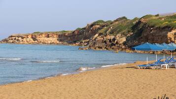 Una spiaggia con acque cristalline, una riva sabbiosa e scogliere rocciose. Si vedono ombrelloni e lettini blu.