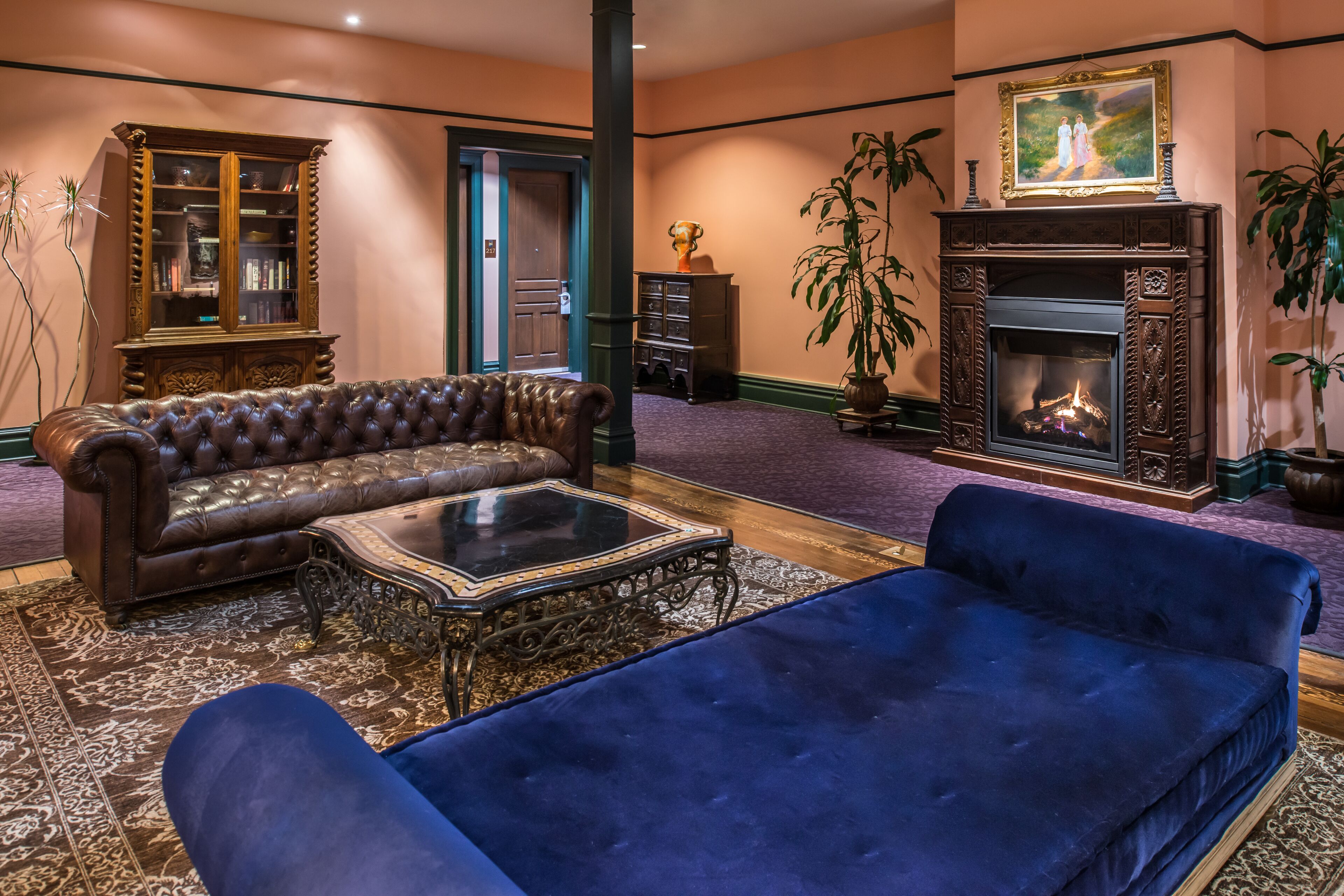 A spacious living room with a brown leather sofa, a blue velvet sofa, a glass coffee table, a fireplace, and a large mirror.