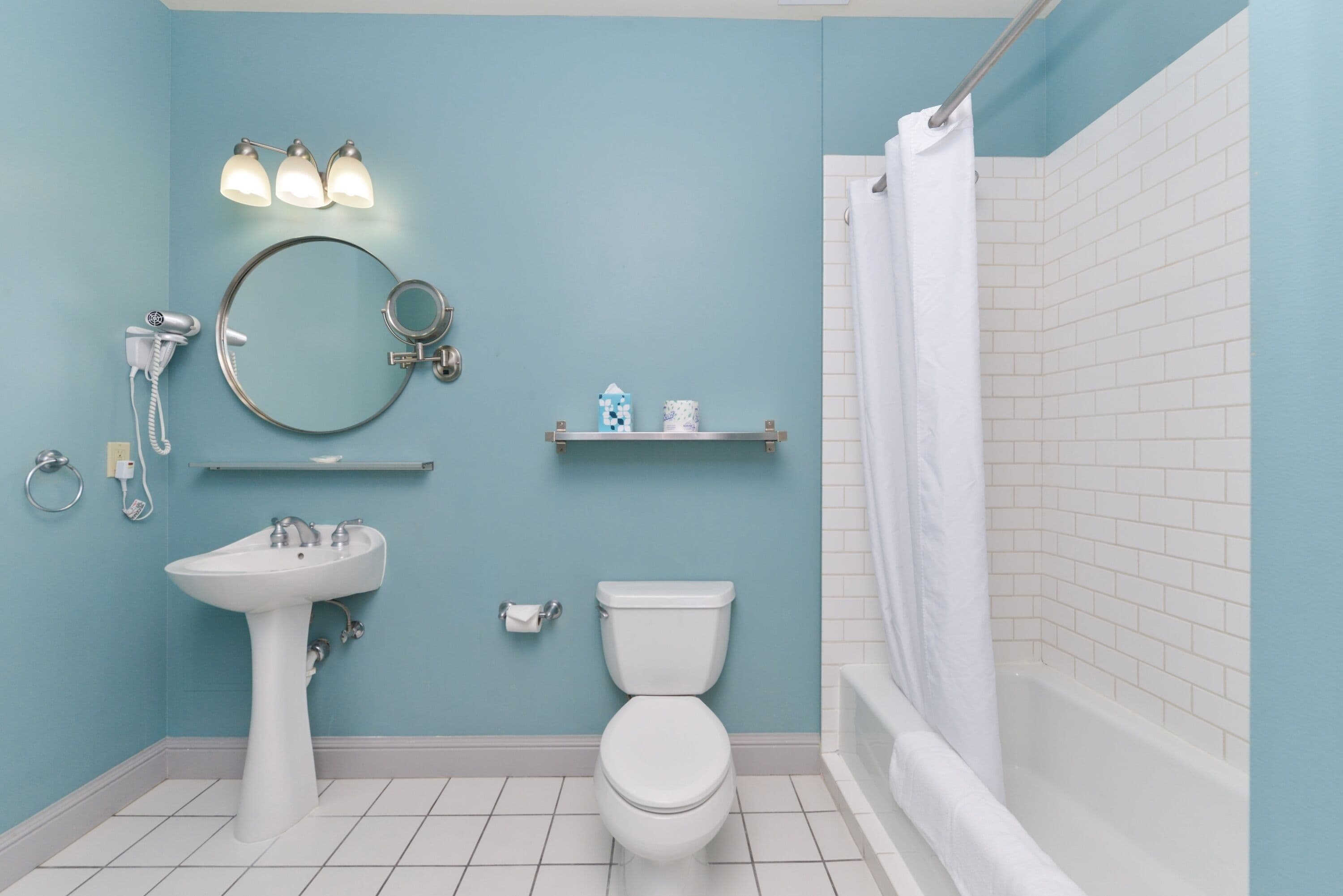 A bathroom with a pedestal sink, a toilet, a shower with a curtain, and a mirror with lights.