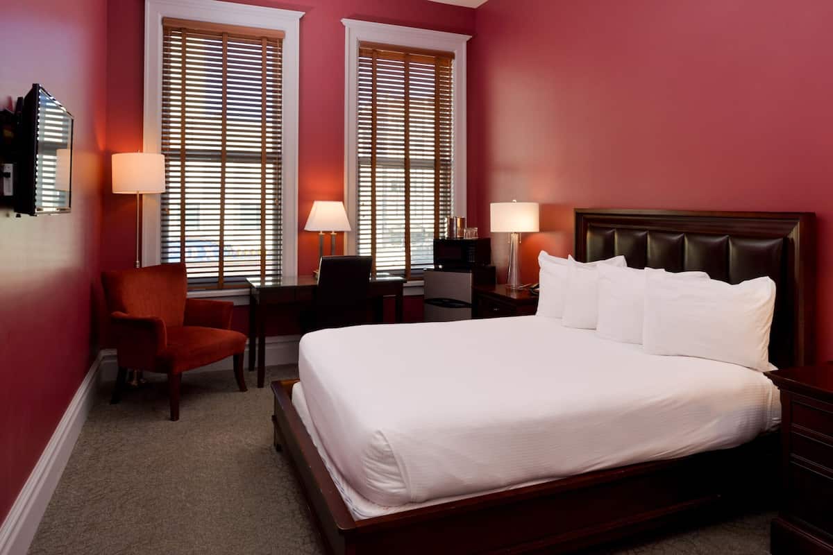 A hotel room with a large bed, two bedside tables, a desk with a chair, and two windows with blinds.