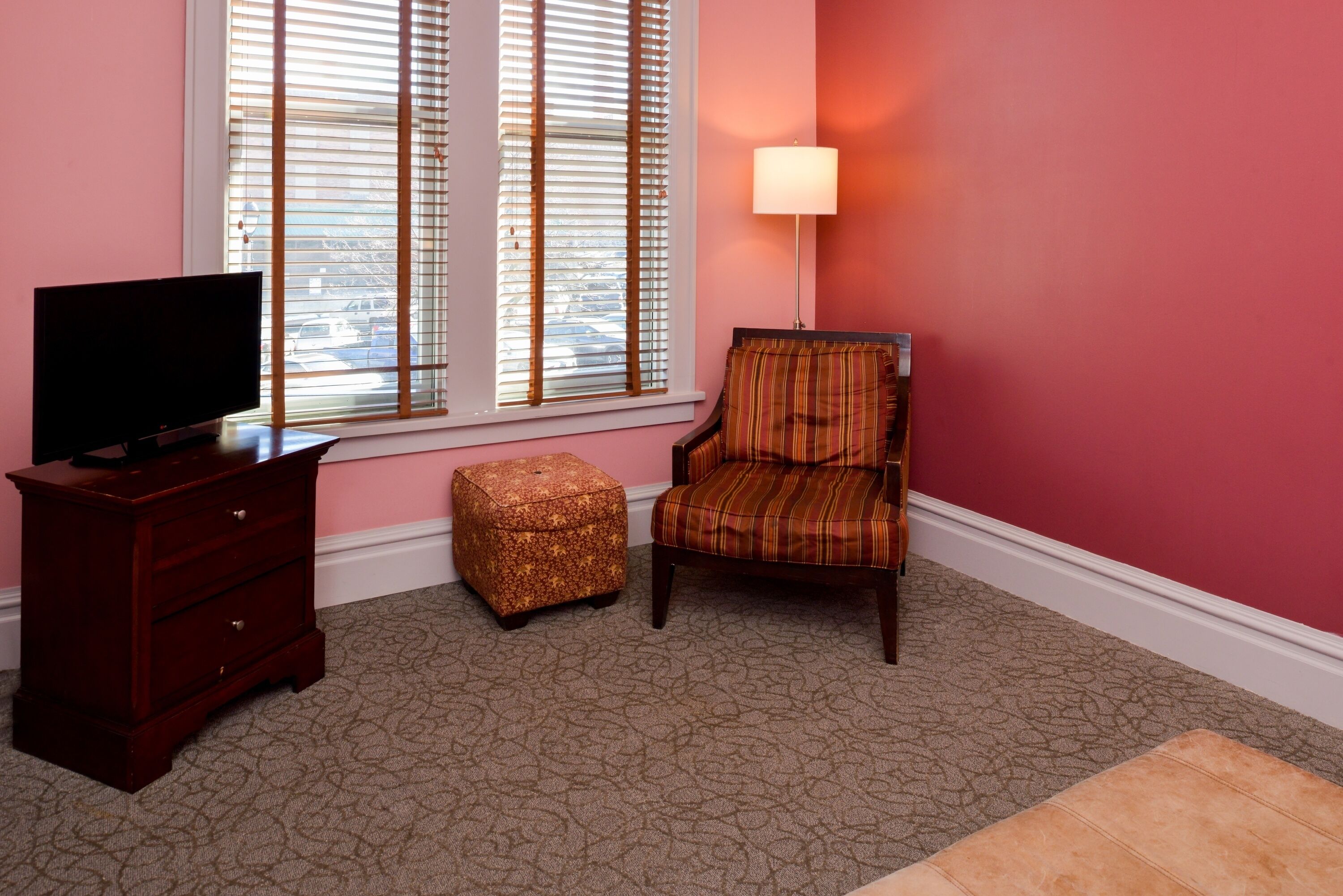 A room with a television on a wooden dresser, a plaid chair, a small ottoman, and a window with blinds.