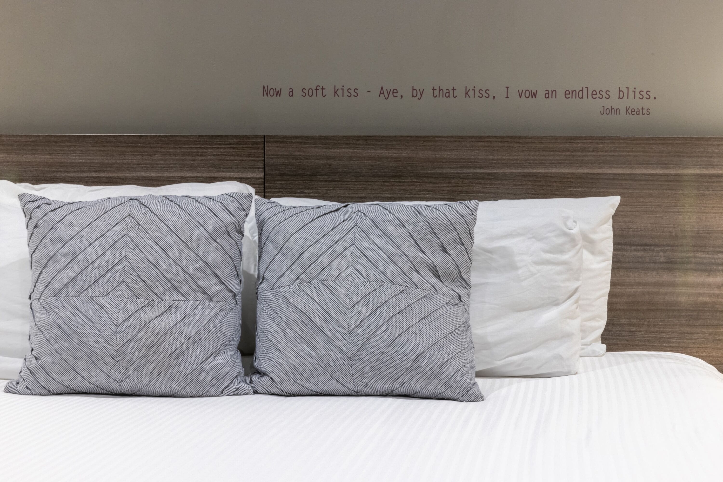 A bed with two patterned pillows and a plain pillow, a wooden headboard, and a quote on the wall.