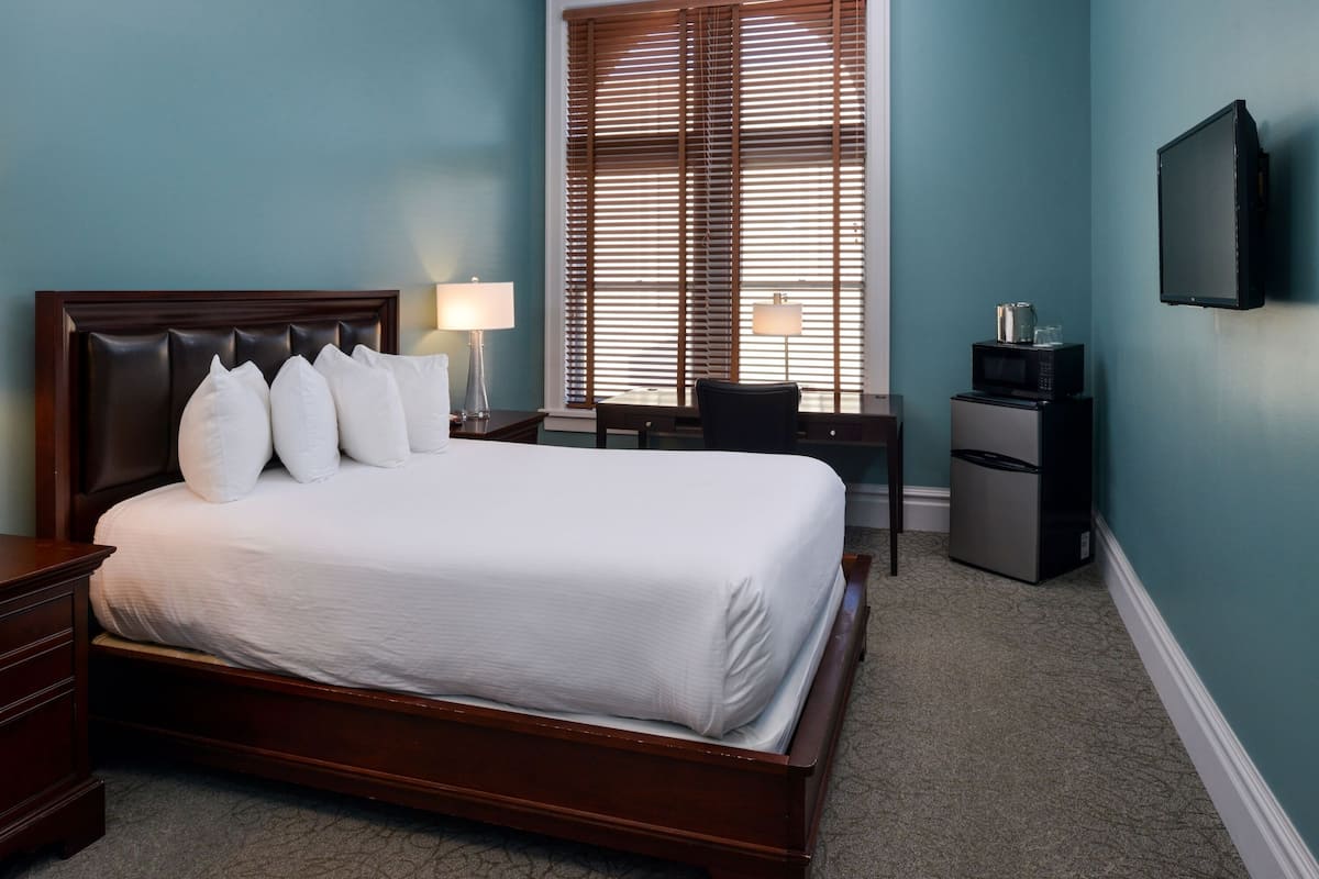 A neatly made bed with white linens, a wooden headboard, and a bedside table with a lamp. A desk with a chair and a microwave are visible in the corner.