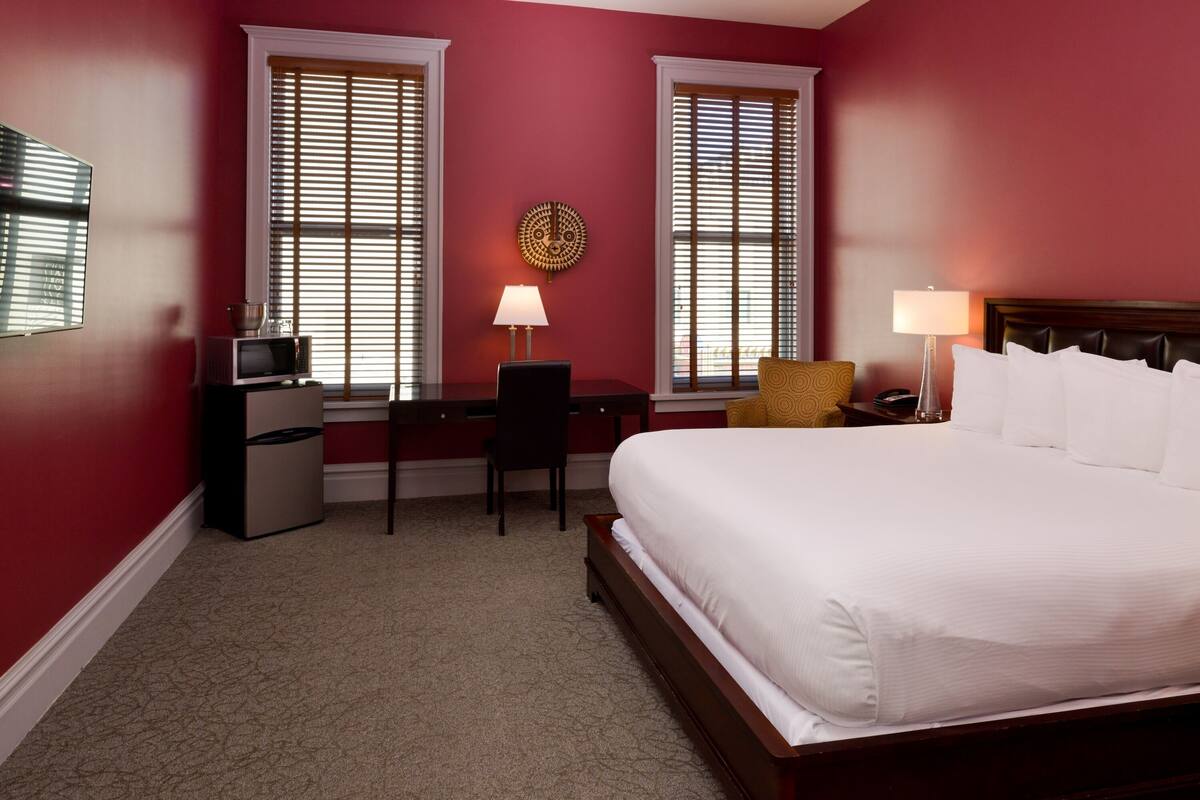 A hotel room with a large bed, a desk with a chair, a small refrigerator, and two windows with blinds.
