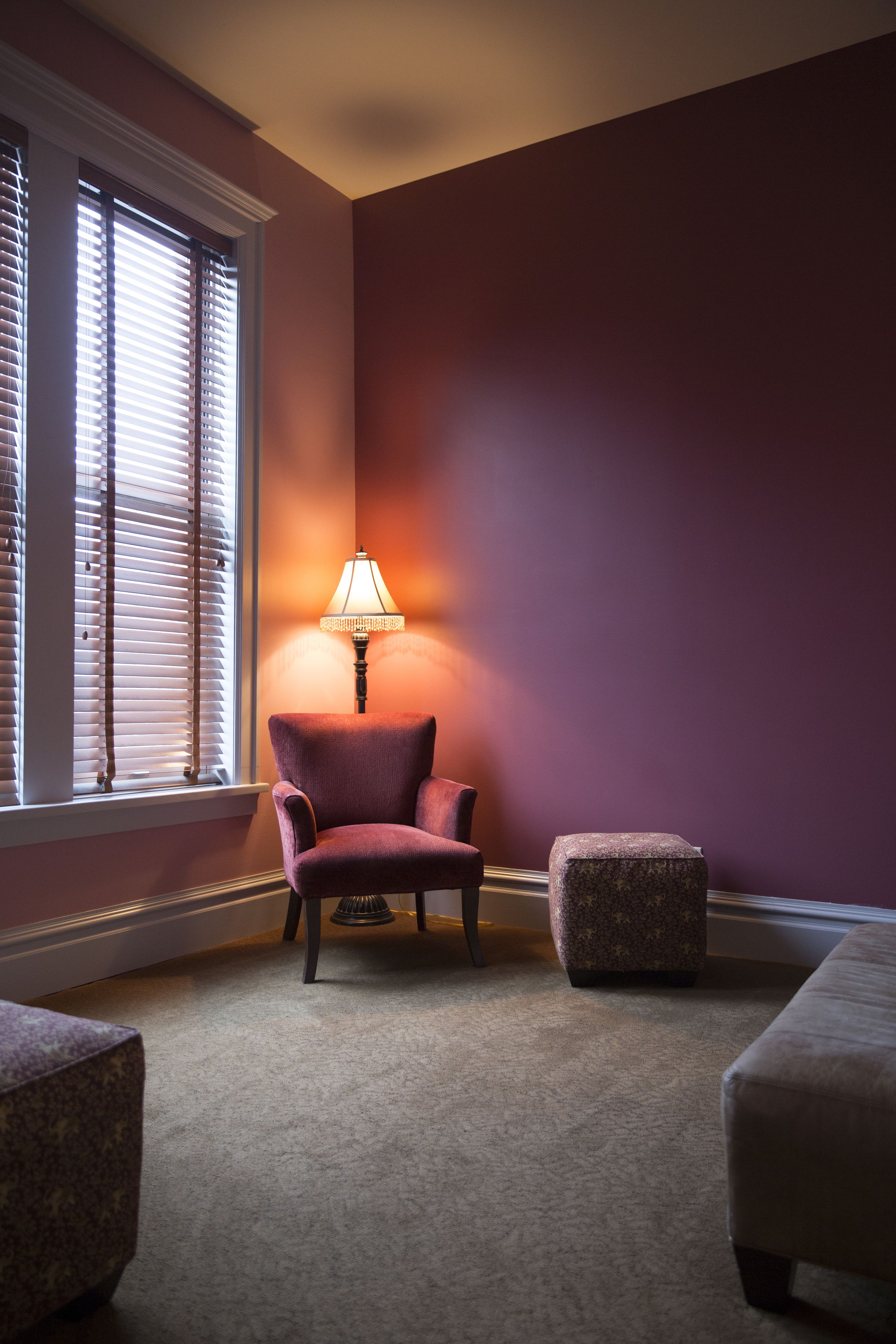 A room with a purple wall, a window with blinds, a chair, a small table, and a lamp.