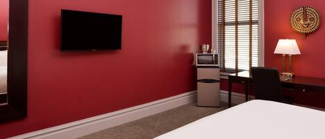 A hotel room with a large bed, a TV mounted on the wall, a desk with a lamp, and a window with blinds.