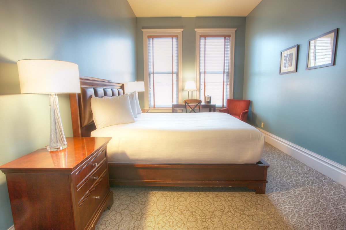 A bedroom with a large bed, a nightstand, a lamp, a chair, and two windows with blinds.