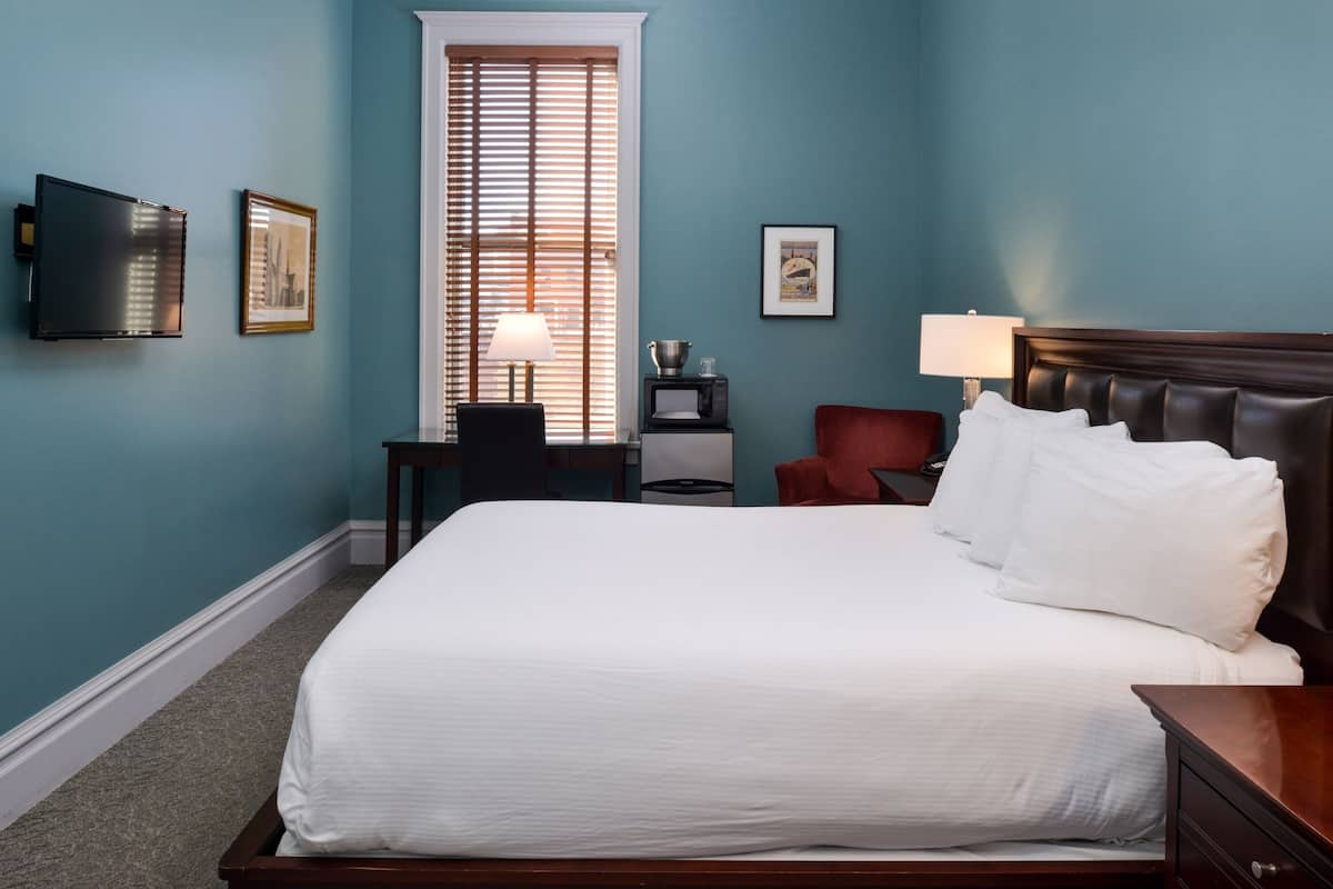 A hotel room with a large bed, a television mounted on the wall, a desk with a chair, and a window with blinds.
