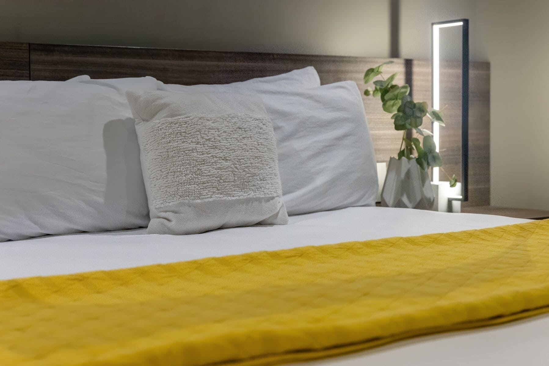 A neatly made bed with white pillows and a yellow blanket, a bedside plant, and a modern bedside table with a mirror.