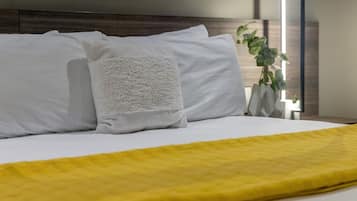 A neatly made bed with white pillows and a yellow blanket, a bedside plant, and a modern bedside table with a mirror.