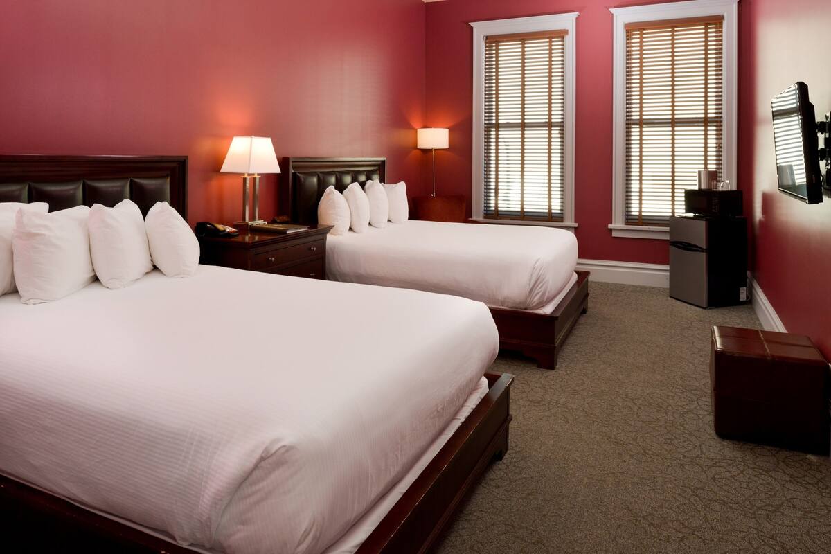 A hotel room with two beds, a nightstand, a lamp, a TV, and a window with blinds.