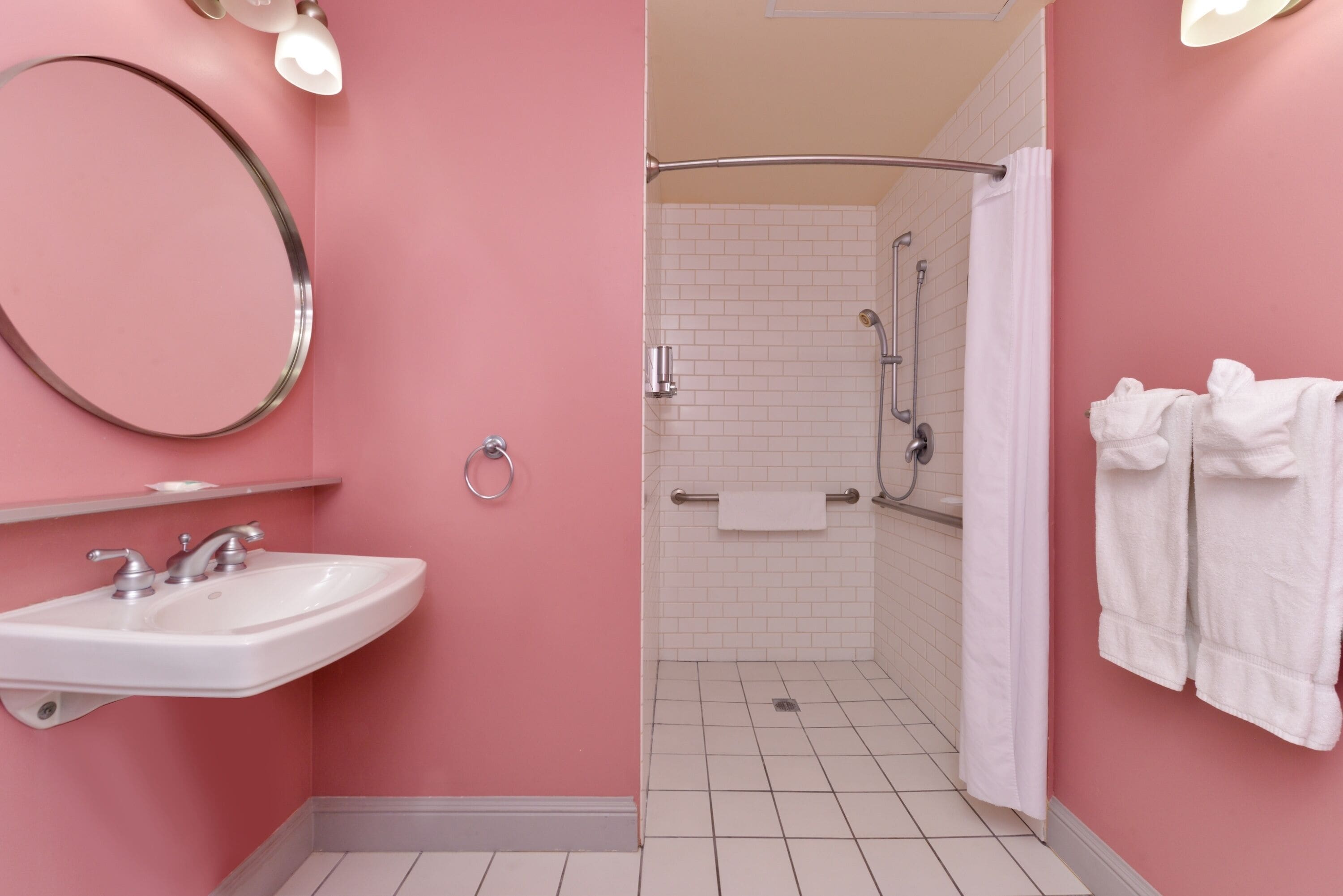 A bathroom with a pink wall, white tiles, a sink with a mirror, a shower with a curtain, and towels on the wall.