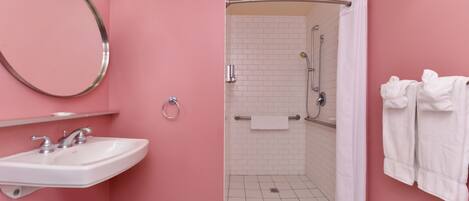 A bathroom with a pink wall, white tiles, a sink with a mirror, a shower with a curtain, and towels on the wall.