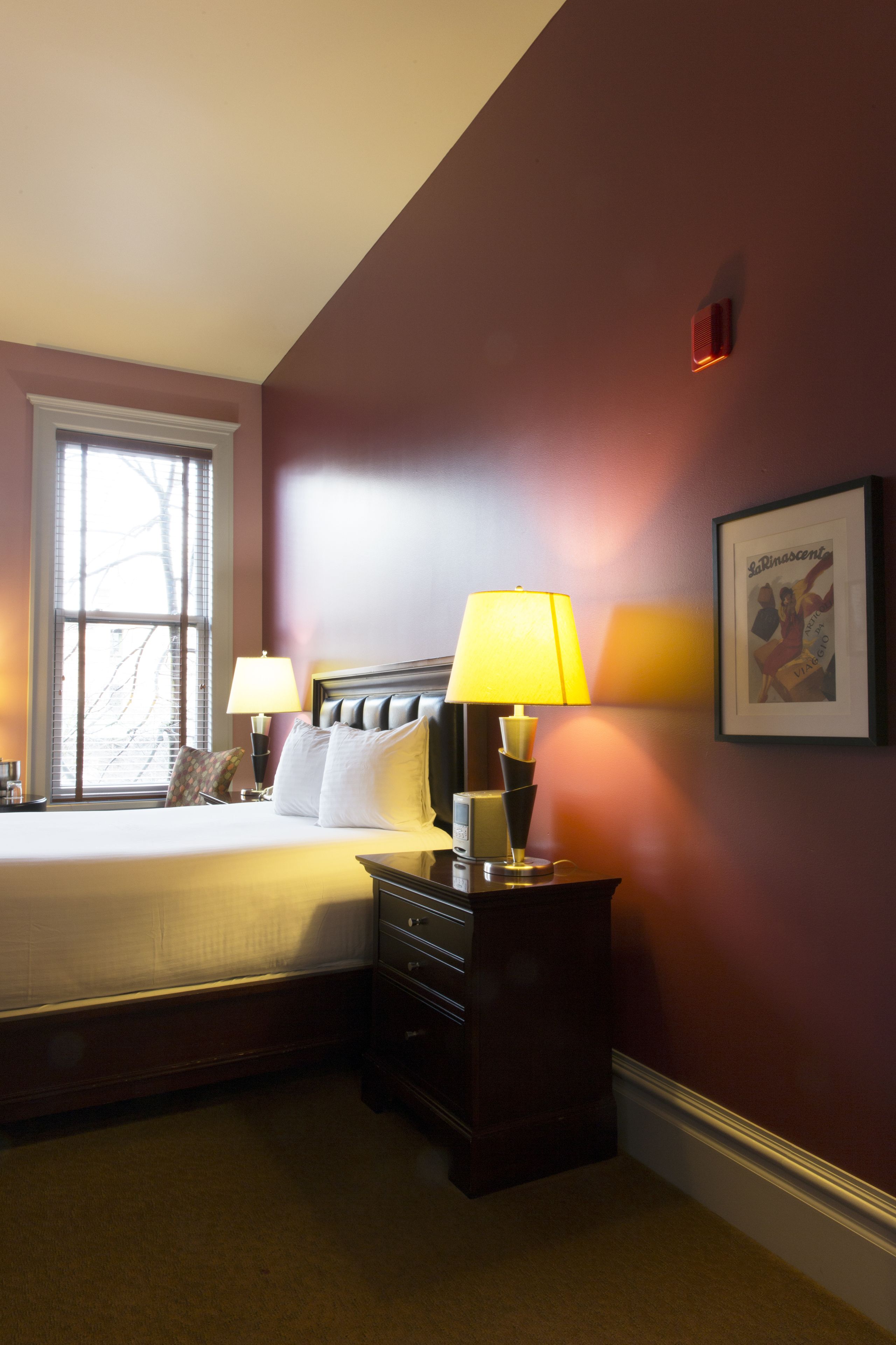 A hotel room with a bed, two bedside lamps, a nightstand, a window with blinds, and a framed picture on the wall.