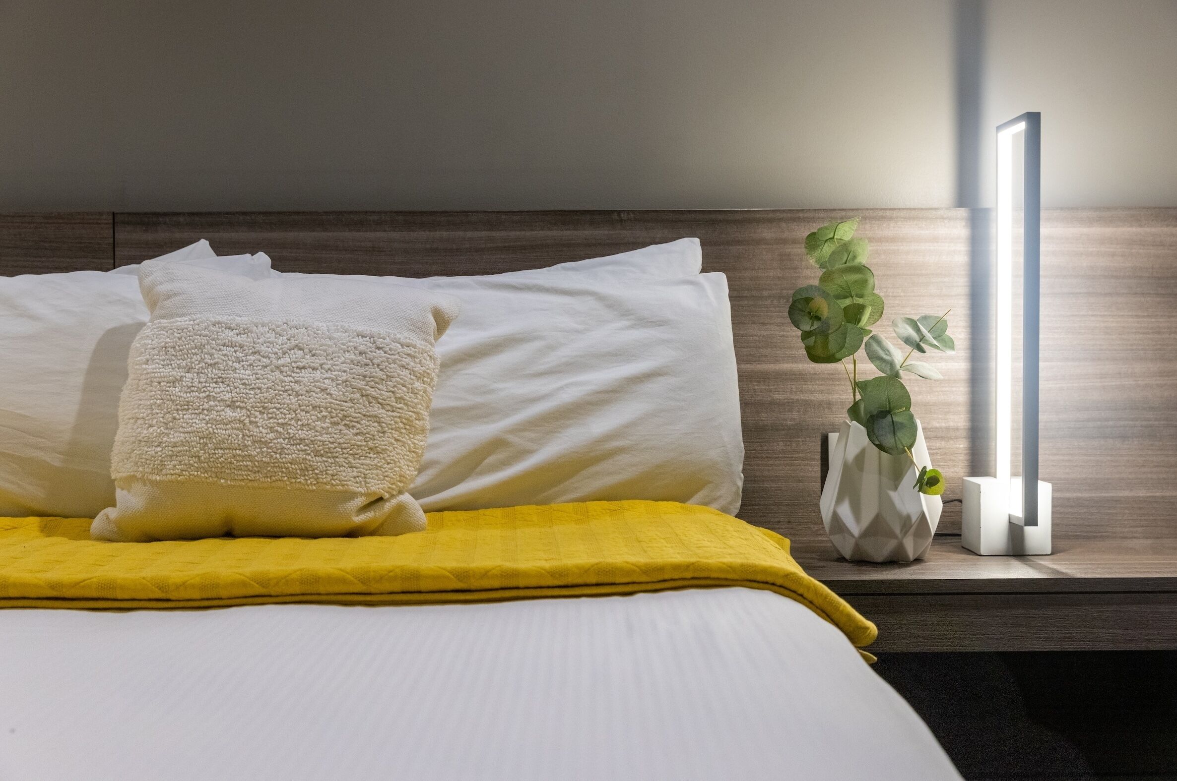 A neatly made bed with white and yellow bedding, a white textured pillow, and a potted plant on a bedside table.