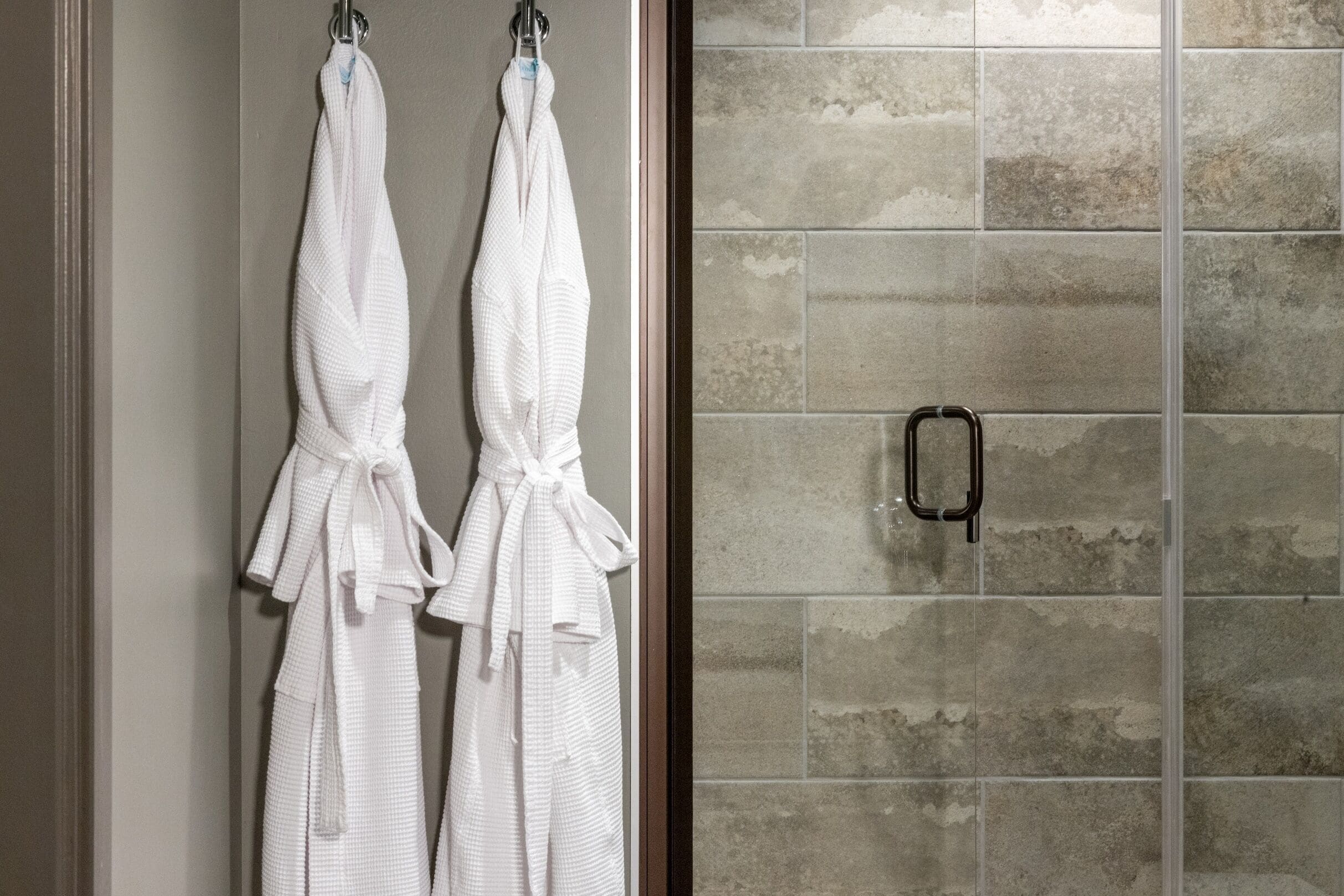 A bathroom with two white towels hanging on hooks, a shower area with a glass door, and a tiled wall.