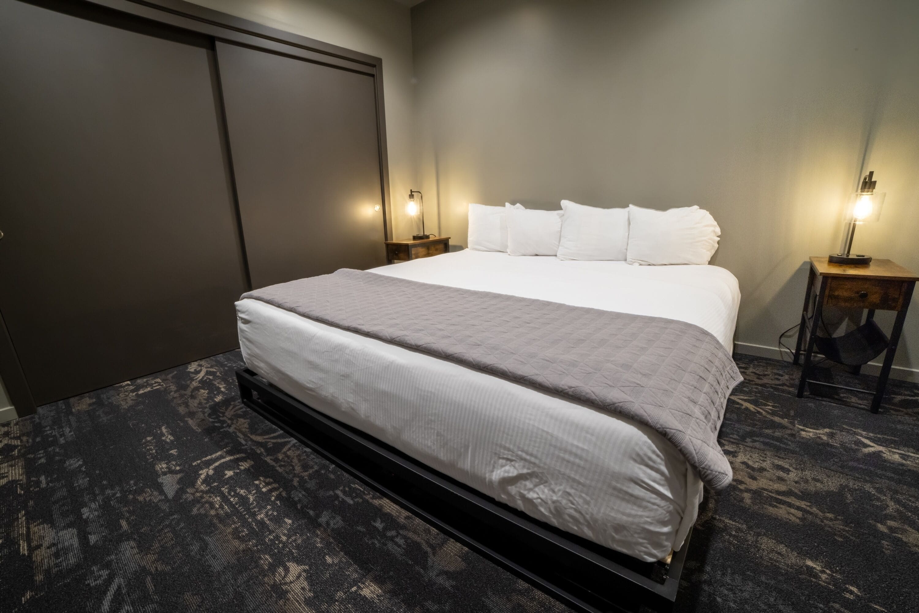 A hotel room with a large bed, two bedside tables with lamps, a wardrobe, and a carpeted floor.