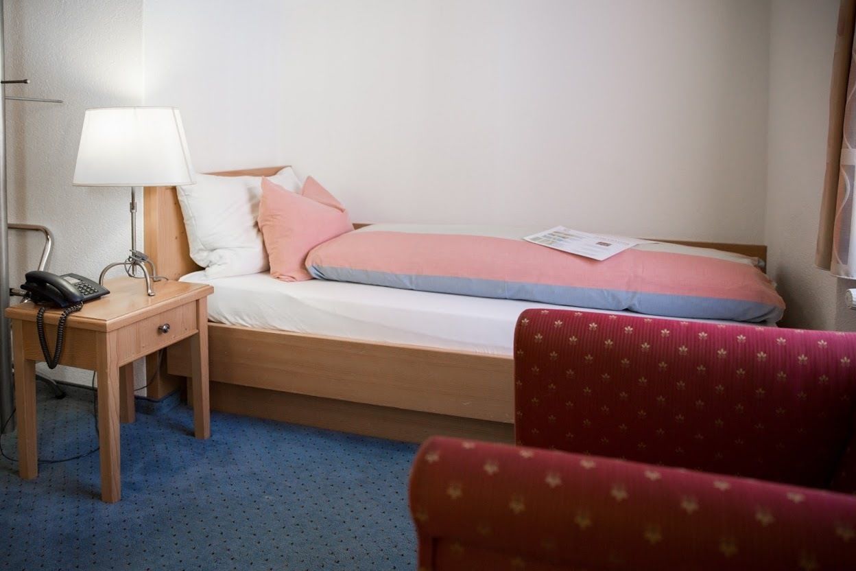 Une chambre d’hôtel avec un lit, une table de chevet avec une lampe, un téléphone et un fauteuil rouge recouvert de tissu.