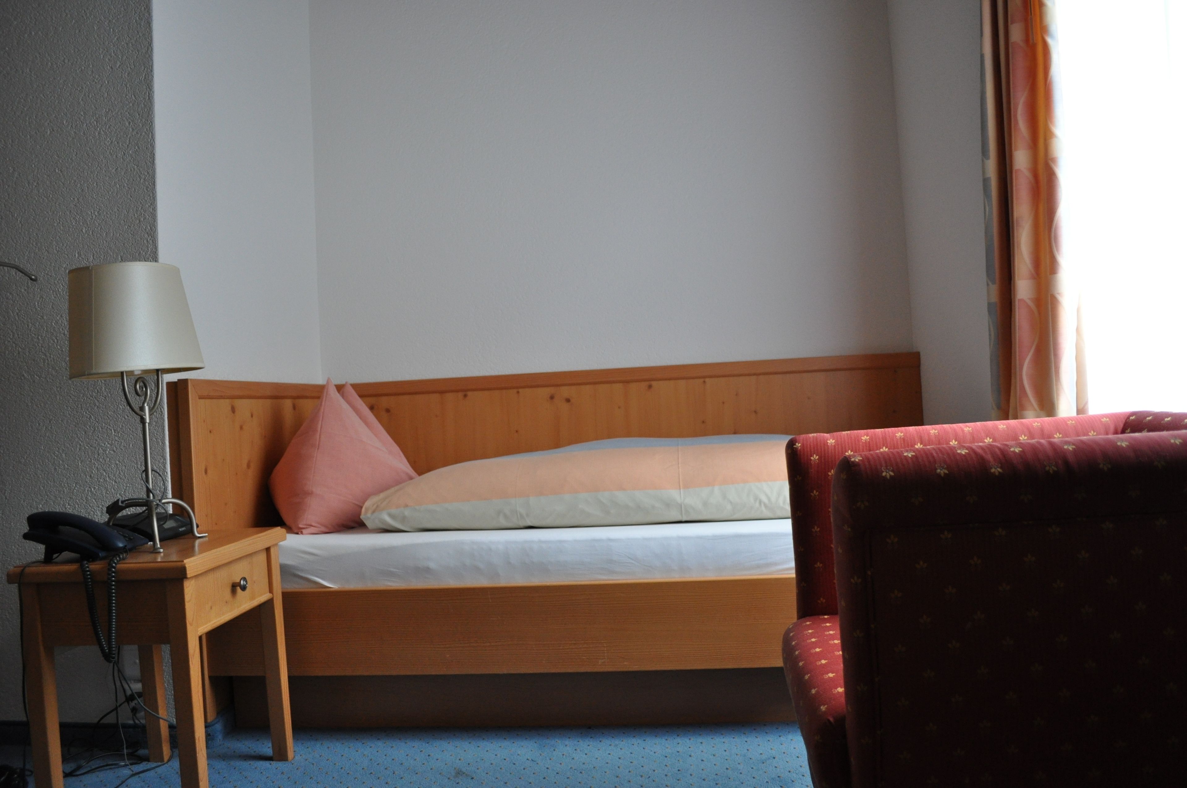 A hotel room with a wooden bed, a bedside table with a lamp, a telephone, and a red upholstered chair.