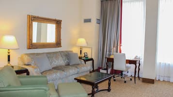 A hotel room with a sofa, armchair, ottoman, a glass-top coffee table, a desk with a chair, a mirror, and a window with curtains.