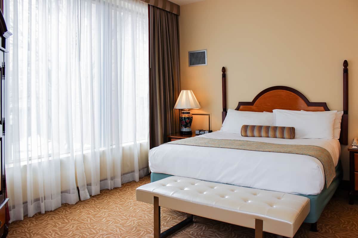 A hotel room with a large bed, a bedside table with a lamp, a bench, and a window with sheer curtains.