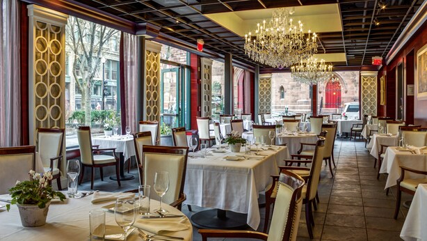 Un restaurant doté de nombreuses tables dressées pour le service, agrémenté de lustres et de grandes fenêtres.