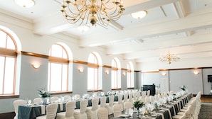 Une salle de banquet avec de longues tables, des chaises blanches, des lustres et de grandes fenêtres.