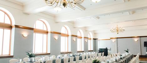 Un salón de banquetes con largas mesas, sillas blancas, candelabros y ventanas grandes.