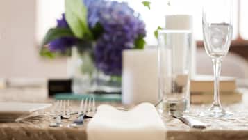 Una mesa dispuesta con una vela blanca, un vaso de agua y una copa de vino, con un jarrón de flores moradas al fondo.
