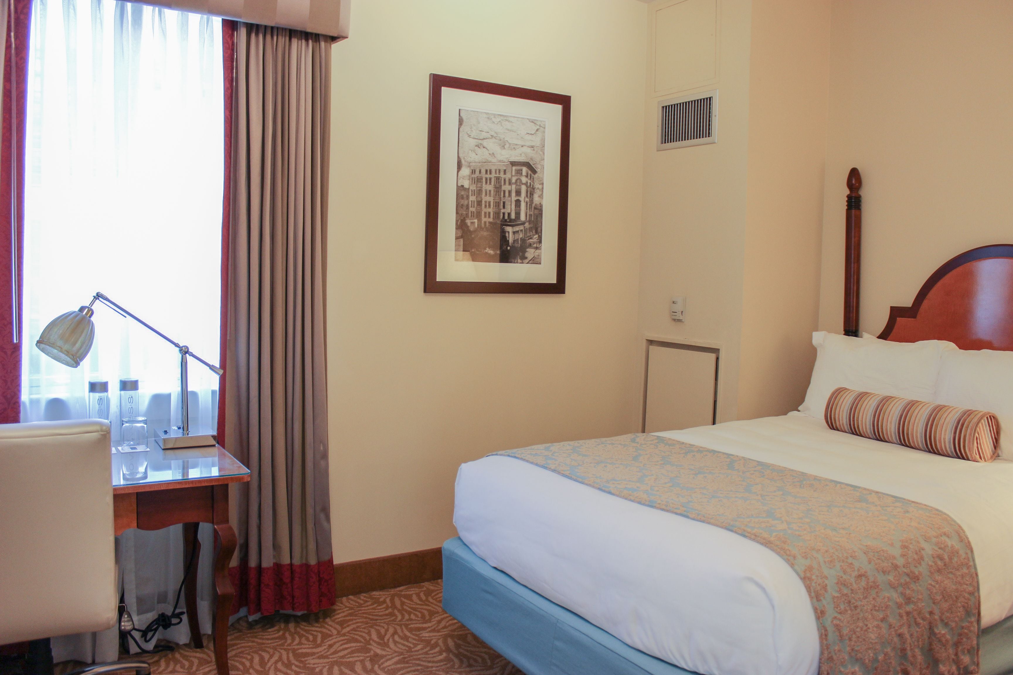 A hotel room with a bed, a desk with a lamp, a framed picture on the wall, and a window with curtains.