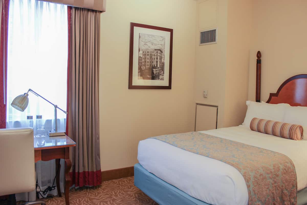 A hotel room with a bed, a desk with a lamp, a framed picture on the wall, and a window with curtains.