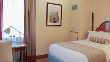 A hotel room with a bed, a desk with a lamp, a framed picture on the wall, and a window with curtains.
