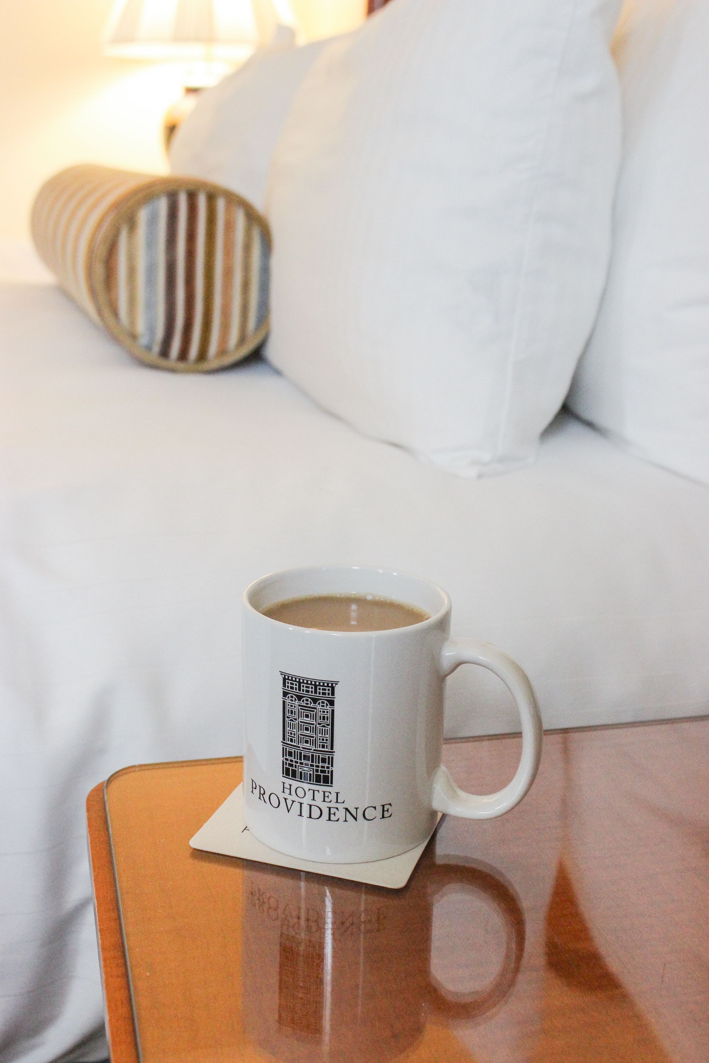 Eine weiße Tasse mit Hotel-Logo auf einem Untersetzer, platziert auf einem Holznachttisch in einem Hotelzimmer.