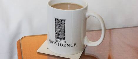 Una taza blanca con el logo de un hotel sobre un posavasos, sobre una mesita de madera en una habitación de hotel.