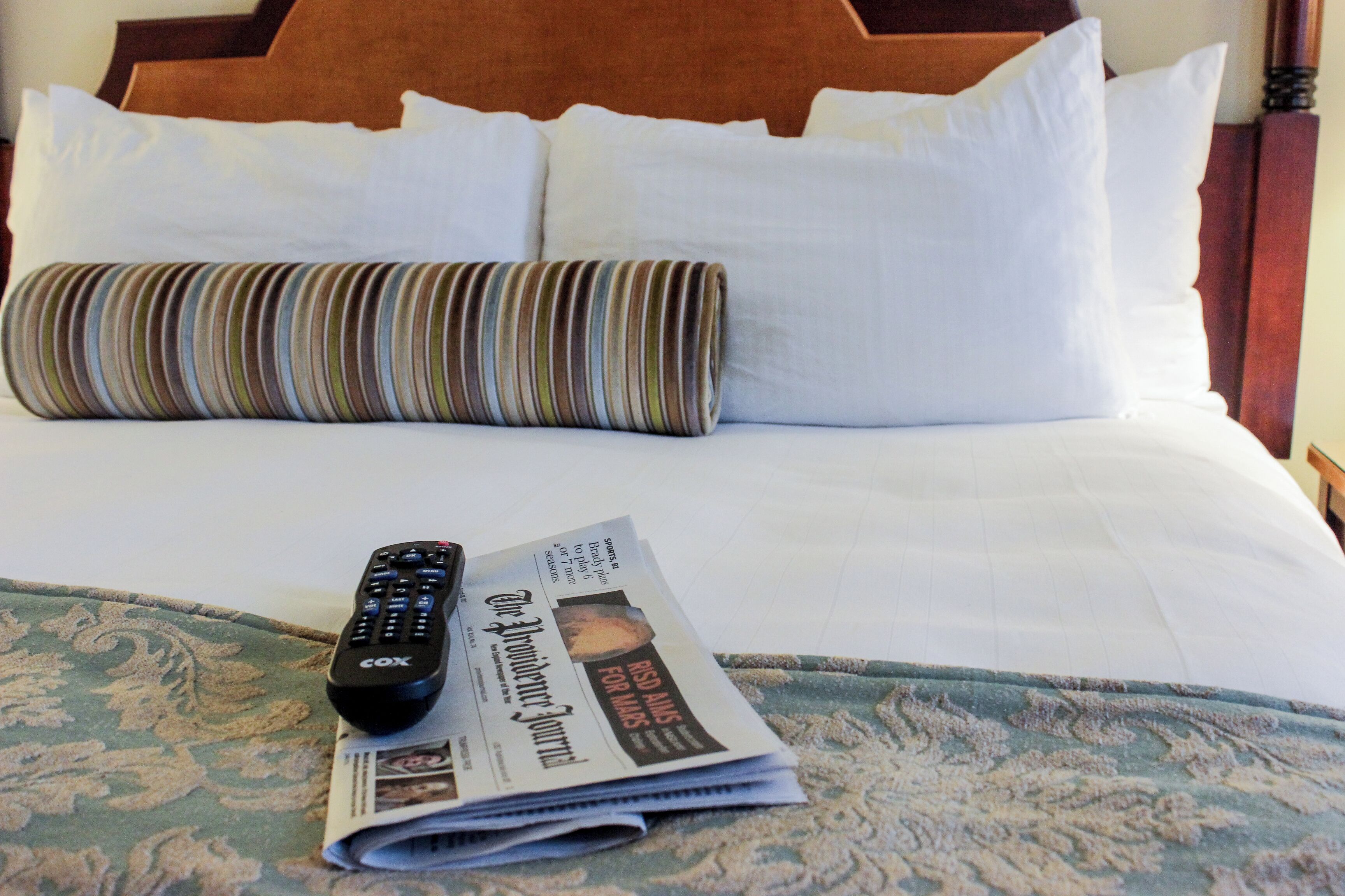 Un lit d’hôtel avec du linge de lit blanc, un oreiller à rayures, une télécommande et un journal plié.