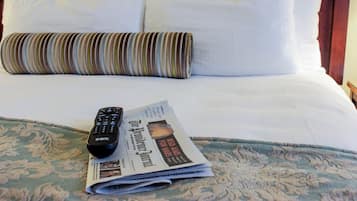 Un lit d’hôtel avec du linge de lit blanc, un oreiller à rayures, une télécommande et un journal plié.