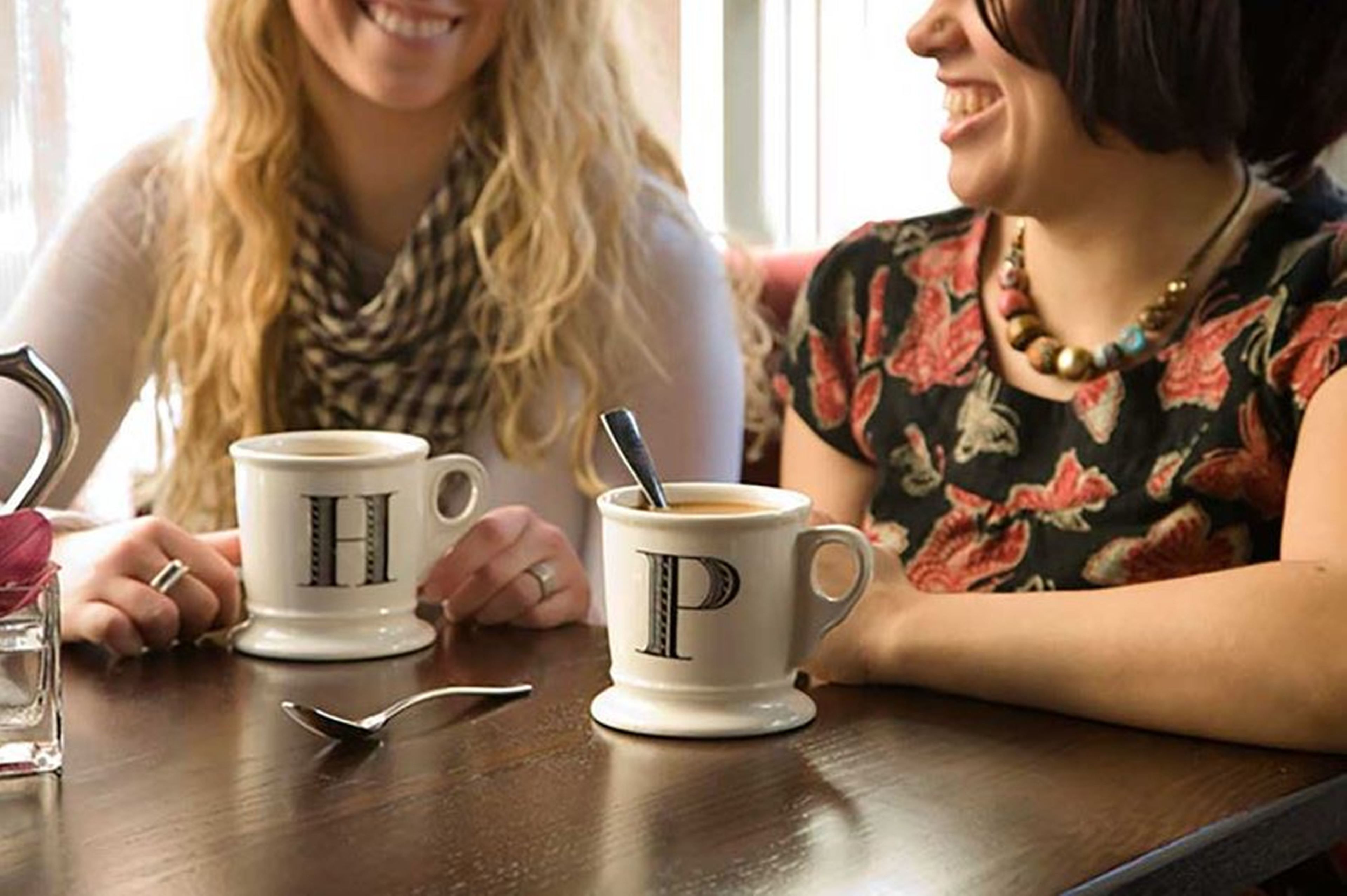 Two people sitting at a table with coffee cups marked with letters, a spoon, and a glass of water.