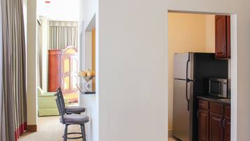 A kitchen with a refrigerator, microwave, and wooden cabinets. There is a chair and a small table with fruit.