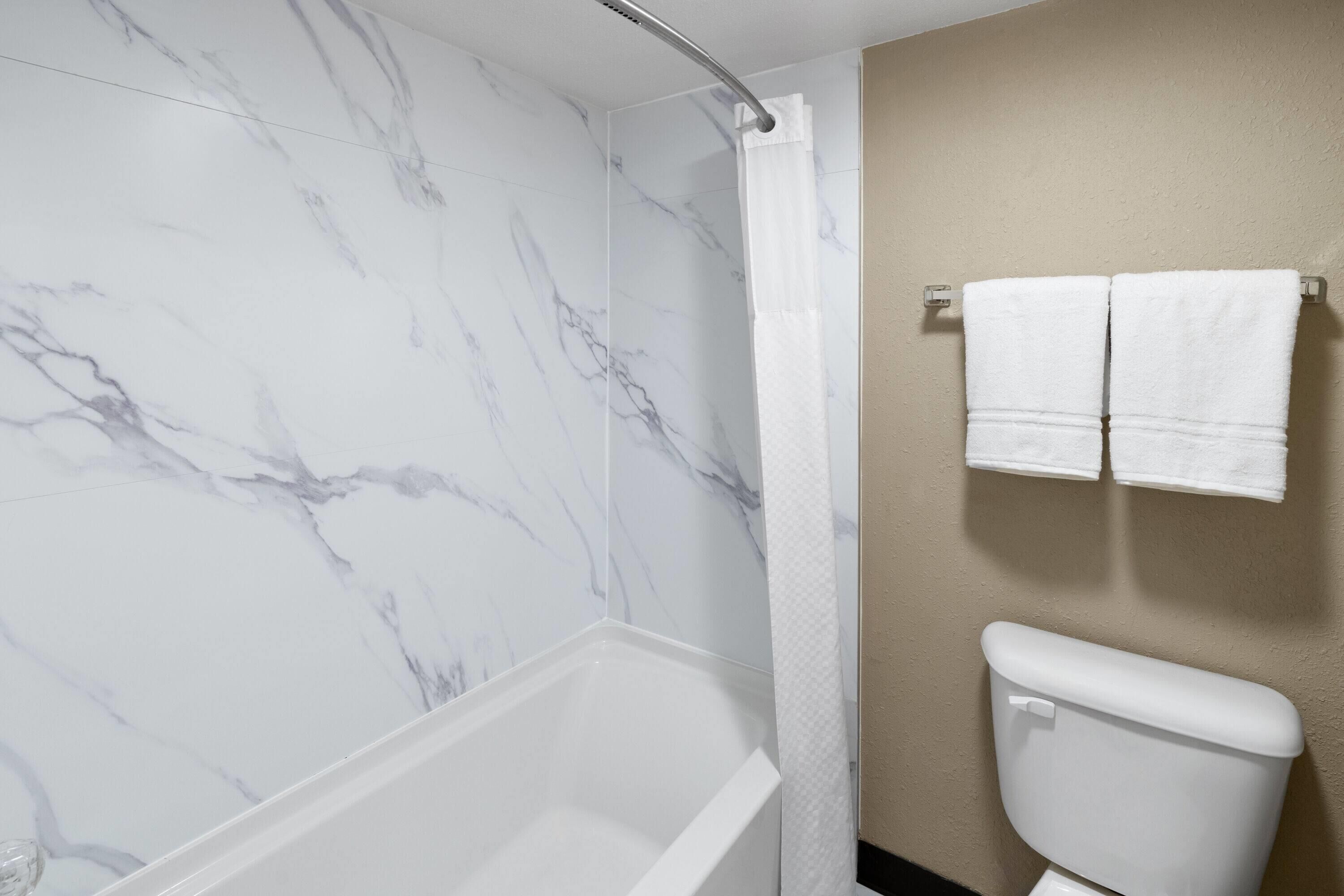 A bathroom with a marble shower wall, a white bathtub, and a toilet with two white towels hanging on the wall.