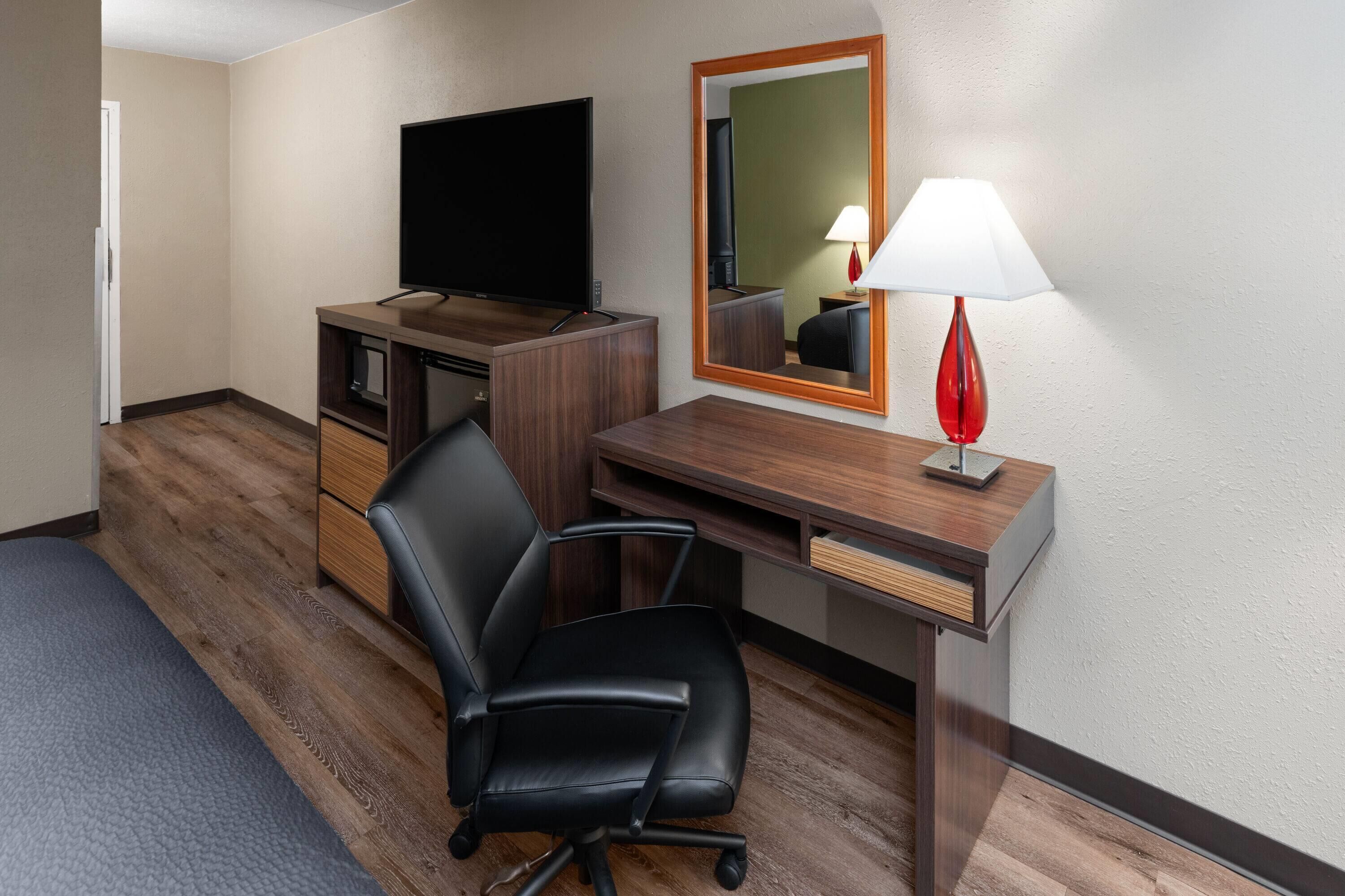 A hotel room with a wooden desk, a black office chair, a flat-screen TV, a mirror, and a red lamp.