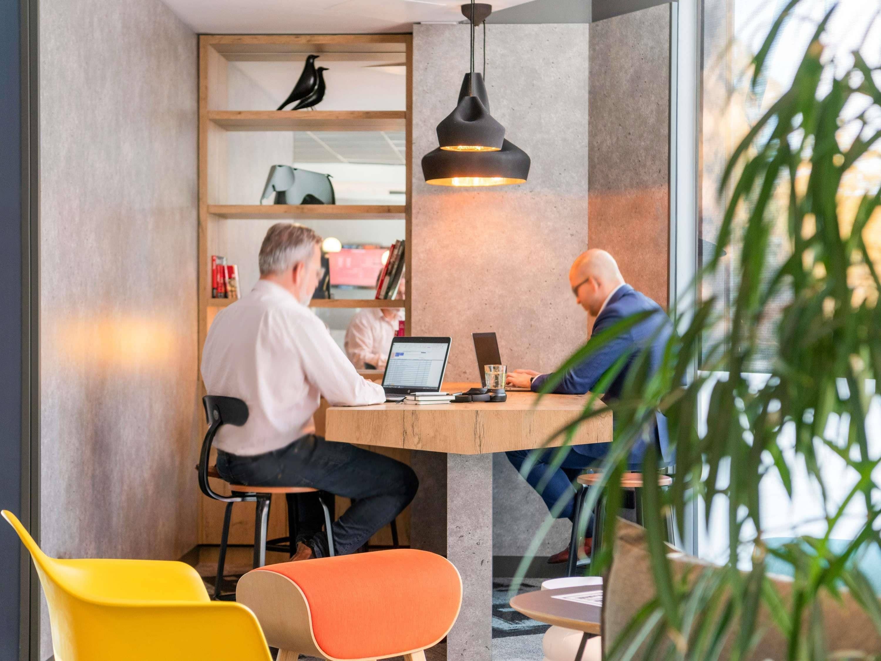 Un ufficio moderno con due persone che lavorano a un tavolo di legno, circondato da sedie colorate e una libreria con oggetti decorativi.