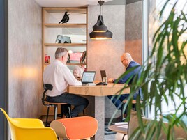 A modern office space with two people working at a wooden table, surrounded by colorful chairs and a bookshelf with decorative items.