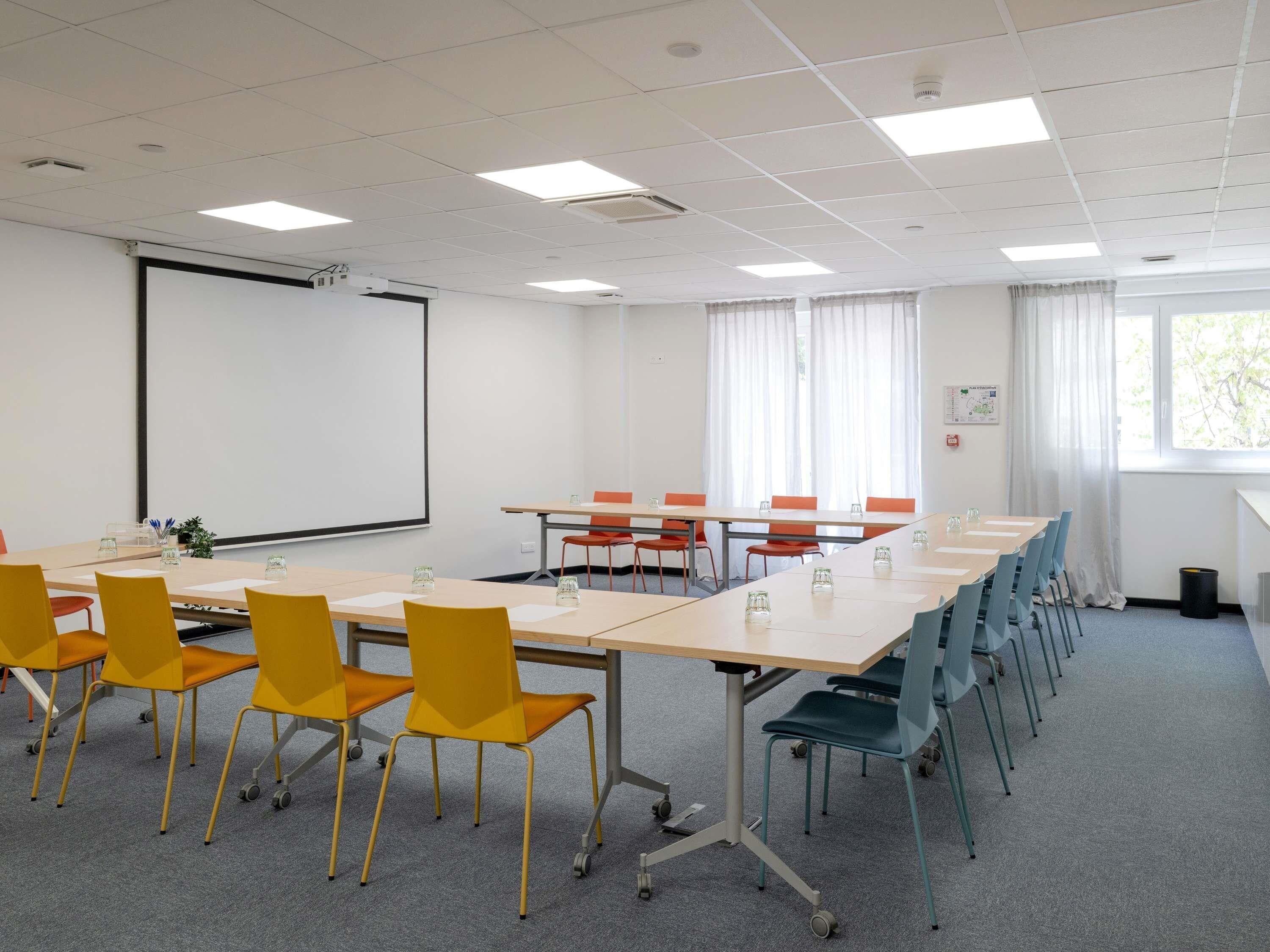 A conference room with a projector screen, tables, chairs, and a window with curtains.