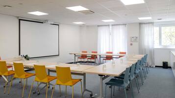 A conference room with a projector screen, tables, chairs, and a window with curtains.