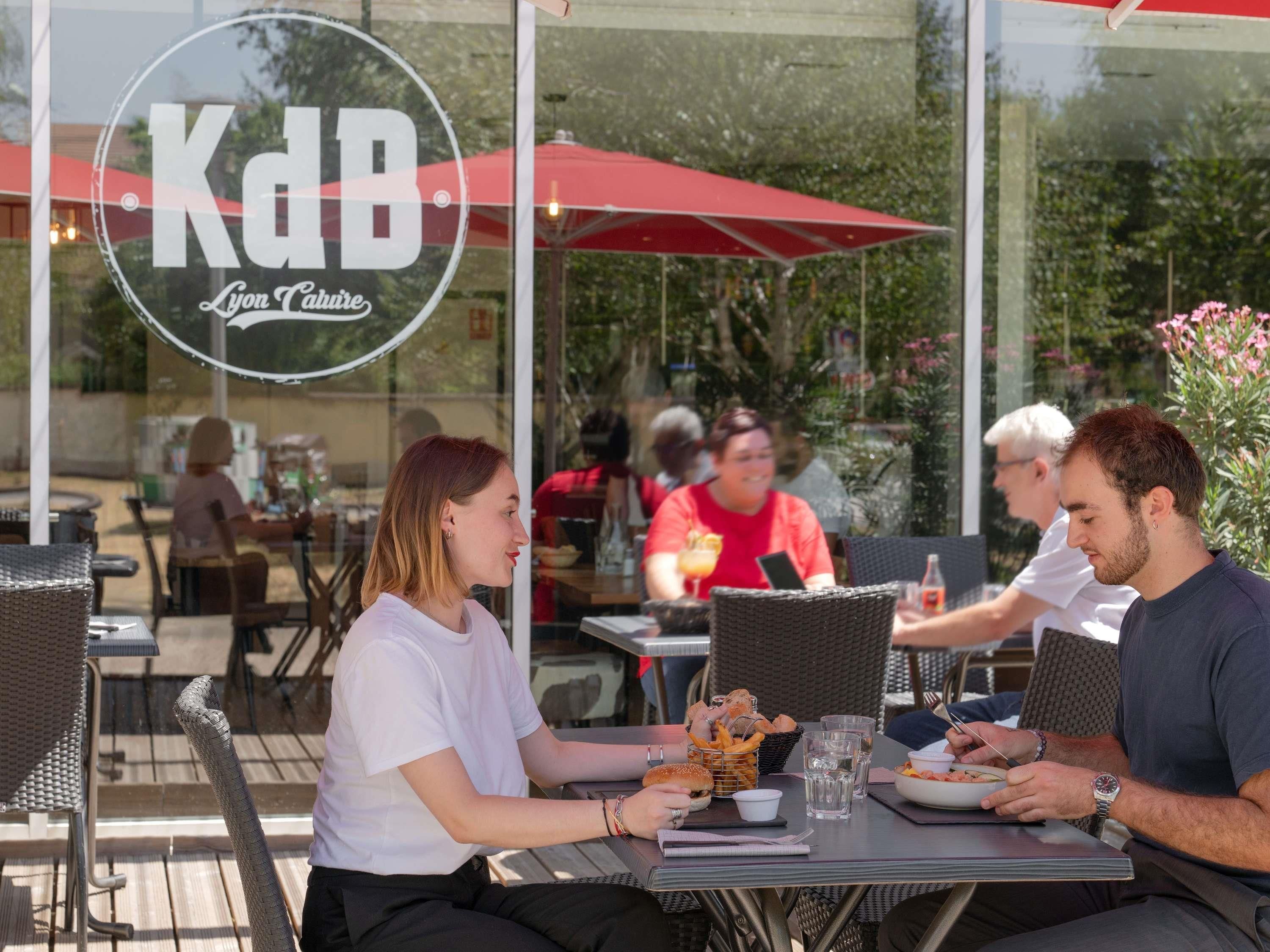 An outdoor dining area with wicker chairs and tables, a red umbrella, and a logo for Kdb Lyon Caillette.