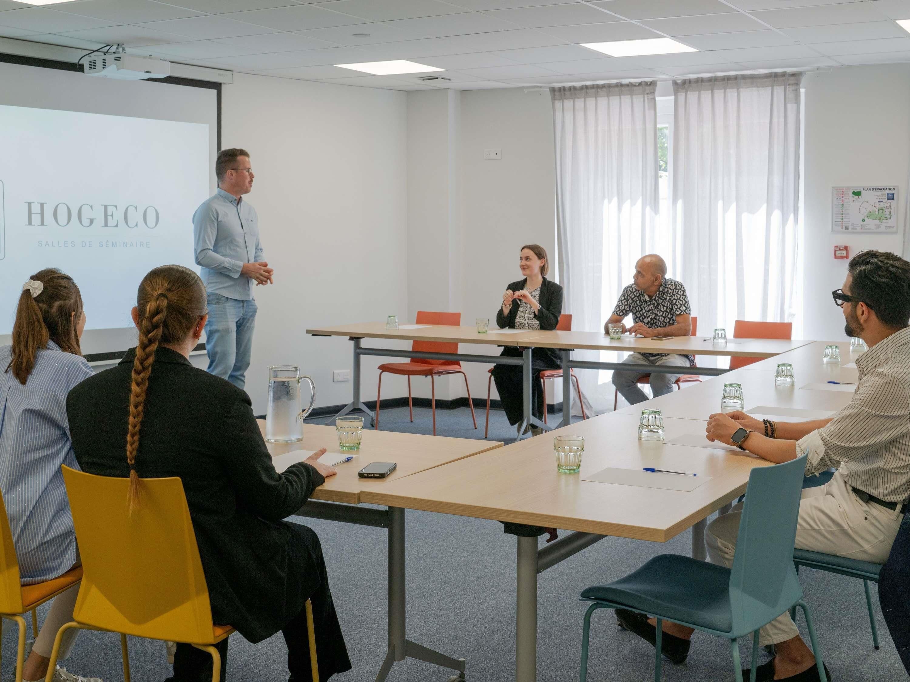 Ein Konferenzraum mit Projektorleinwand, einer Präsentation und Teilnehmern, die um einen Tisch sitzen.