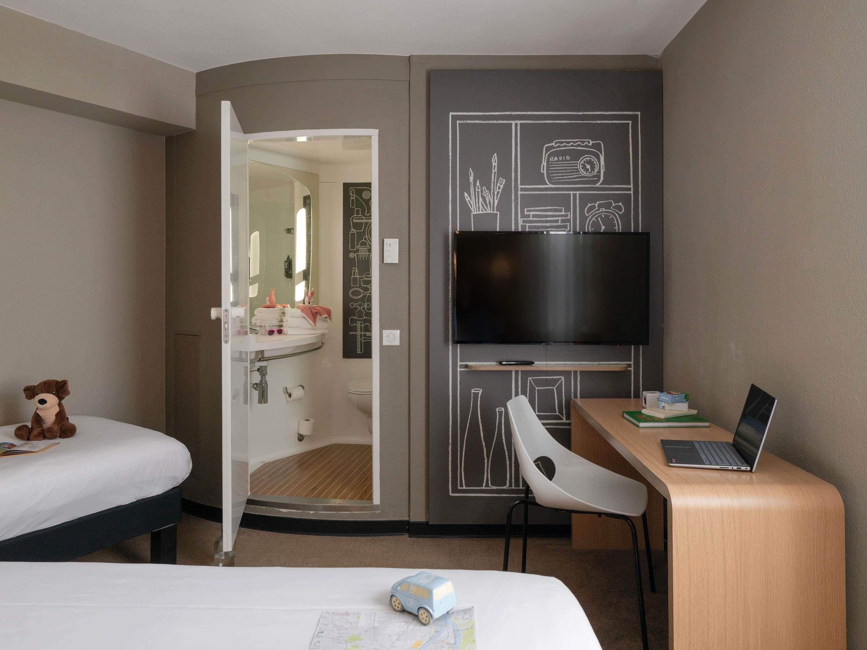 A hotel room with a bed, a desk with a laptop, a TV, and a teddy bear on the bed.