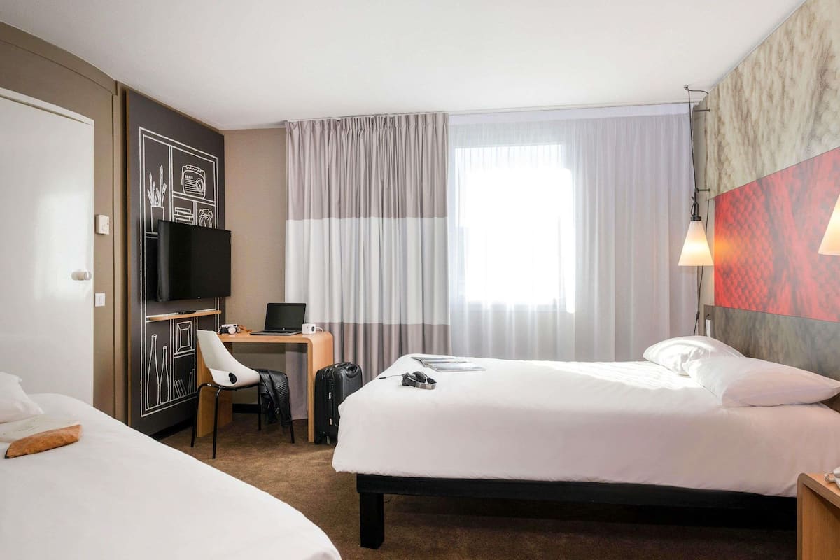 A hotel room with two beds, a desk with a laptop, a TV, and a large window with curtains.